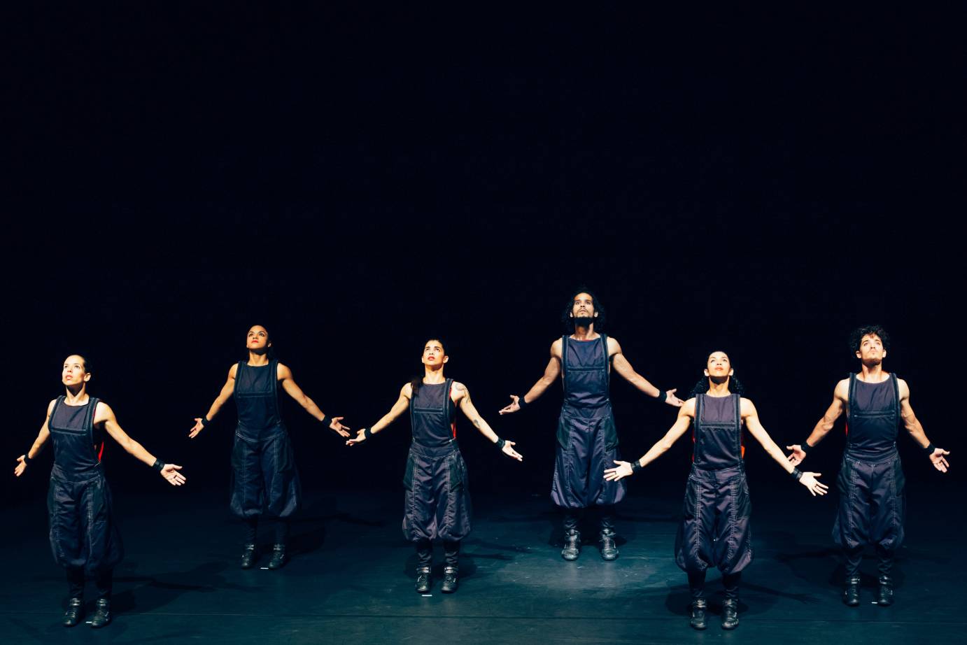 Six dancers extends their arms to the side while looking tough